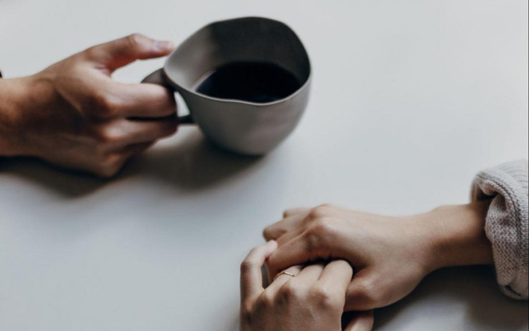 two people's hands on a table, one holding a cup, the other with clasped hands